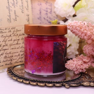 Pu&ograve; includere: Un piccolo barattolo di vetro con un coperchio di rame riempito di una candela rosa con glitter rosso e viola. Il barattolo &egrave; appoggiato su un vassoio decorativo, ornato e dorato.