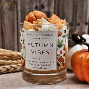 Könnte beinhalten: Eine Sojawachskerze im Glas, verziert mit wachsartigen Dekorationen in Form von Kürbissen und Blättern. Das Etikett trägt die Aufschrift East Light Candles, Autumn Vibes, Pecan Pumpkin Pie Soy Wax Candle, made with Love, mit Herbstlaubmotiven.