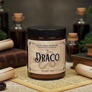Könnte beinhalten: Eine braune Glas-Kerze im Glas mit schwarzem Deckel. Das Etikett trägt die Aufschrift "Draco" mit "Sweet Apple • Amber" darunter. Die Kerze ist von alten Büchern, Schriftrollen und Apothekerflaschen umgeben, was ein mystisches Thema erzeugt.