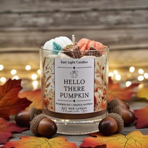 Könnte beinhalten: Ein klares Glas-Kerzenglas mit einem weißen Etikett mit der Aufschrift "Hello There Pumpkin". Die Kerze ist mit dekorativen Wachsformen und einem Docht versehen. Die Kerze steht auf einem Bett aus Herbstblättern und Eicheln.