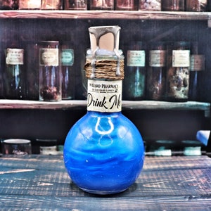 Peut inclure: Une bouteille en verre bleu avec un bouchon argenté et brun. La bouteille est remplie d'un liquide bleu et porte une étiquette sur laquelle est inscrit "Wizard Pharmacy by East Eight Candles" et "Drink Me".