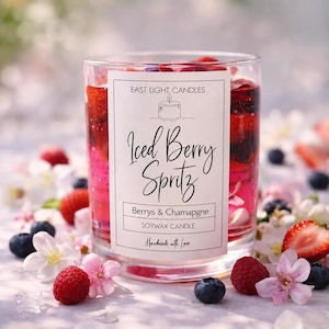 May include: A clear glass candle filled with pink wax, red berries, and dark fruit pieces. The white label reads East Light Candles, Iced Berry Spritz, Berrys & Chamapgne, Soywax Candle, and Handmade with Love, featuring a line drawing of a candle.