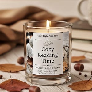Könnte beinhalten: Eine brennende Sojawachskerze in einem durchsichtigen Glas. Das Etikett lautet "Cozy Reading Time" mit dem Duft "Pumpkin Hot Chocolate". Die Kerze ist von Herbstblättern und Nüssen umgeben.
