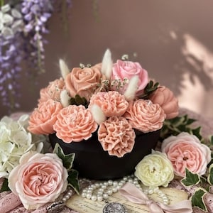 Può includere: Una composizione floreale con rose e garofani color pesca e rosa in una ciotola nera. La composizione è circondata da perle, un ciondolo vintage e un nastro rosa. Lo sfondo comprende un tessuto di pizzo rosa e fiori viola.