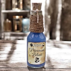 Puede incluir: Una pequeña botella de vidrio llena de líquido azul, etiquetada como "Mermaid Blood". La botella está envuelta con cuerda y tiene un tapón de corcho con un sello de cera. La etiqueta presenta una ilustración de sirena y el texto "Wizard Pharmacy by East Light Candles".