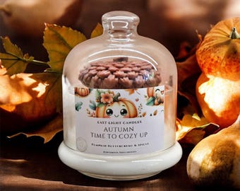 Könnte beinhalten: Eine Glashaubenkerze mit einem kürbisförmigen Aufsatz, die auf einem weißen Sockel steht. Die Kerze hat ein Etikett mit der Aufschrift "Autumn Time to Cozy Up" mit Kürbisillustrationen. Der Hintergrund zeigt Kürbisse und Herbstblätter.