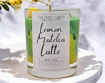 May include: A clear glass candle with a layered green and yellow wax interior. The candle has a label that reads "Lemon Matcha Latte" with the words "Matcha - Lemon" and "Handmade with Love" below. Yellow flowers are in the background.