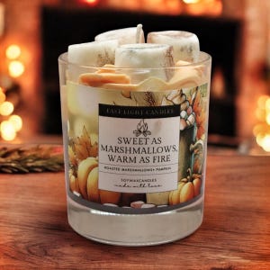 May include: A glass candle with a label that reads "Sweet as Marshmallows, Warm as Fire." The candle has a design of pumpkins and autumn leaves. The candle is filled with white and tan wax cubes.