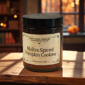 May include: A brown glass jar with a black lid, labeled "Mollys Spiced Pumpkin Cookies" from East Light Candles. The label has a vintage design with the words "Pumpkin Spice & Cookies" at the bottom. The candle sits on a wooden surface.