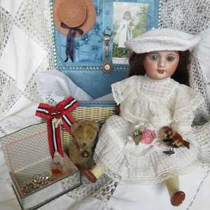 Puede incluir: Una muñeca de porcelana blanca con un vestido de encaje blanco y un sombrero de ganchillo blanco. La muñeca sostiene un pequeño pájaro y una flor. La muñeca está sentada en una cesta de mimbre con un oso de peluche a su lado. La muñeca está rodeada de encaje blanco.