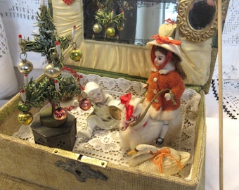 Puede incluir: Una caja de exhibición vintage con temática navideña con un árbol en miniatura, adornos y muñecas. La caja está forrada con tela color crema y presenta un pequeño espejo y una decoración en forma de corazón. Las muñecas visten atuendos festivos.