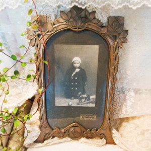 May include: A vintage bronze picture frame holding a monochrome photograph of a child. The frame features a decorative floral and leaf pattern. The child in the photo is wearing a hat and coat, with a toy horse and ball.