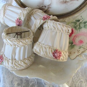 May include: Four ivory lampshades with ruffled edges and pink satin bows. The lampshades are displayed on a white porcelain tray.
