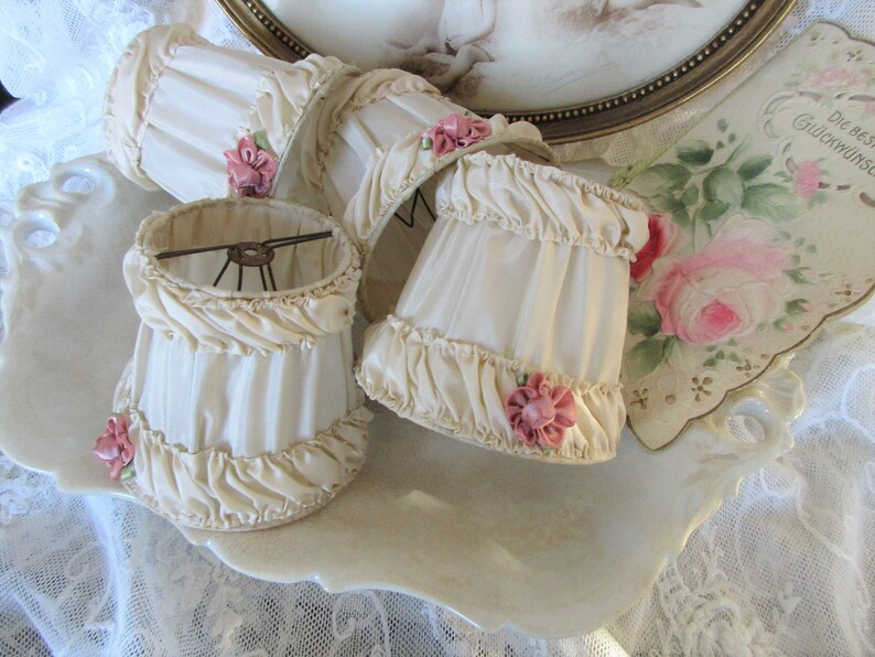 May include: Four ivory lampshades with ruffled edges and pink satin bows. The lampshades are displayed on a white porcelain tray.