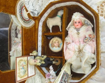 Puede incluir: Un expositor vintage forrado de terciopelo con múltiples compartimentos. Contiene una muñeca con un vestido rosa, una figura de cordero, un retrato y una pequeña muñeca con un atuendo verde. El expositor está adornado con encaje y tiene un espejo.