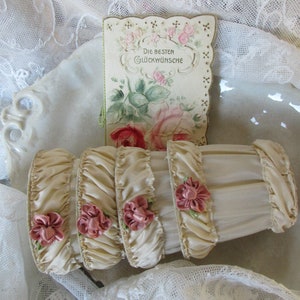 May include: Four ivory satin ribbon ruffles with pink satin flowers and green leaves. The ruffles are arranged in a stack and are sitting on a white lace tablecloth. A vintage card with a floral design and the text "Die Besten Gl&uuml;ckw&uuml;nsche" is behind the ruffles.