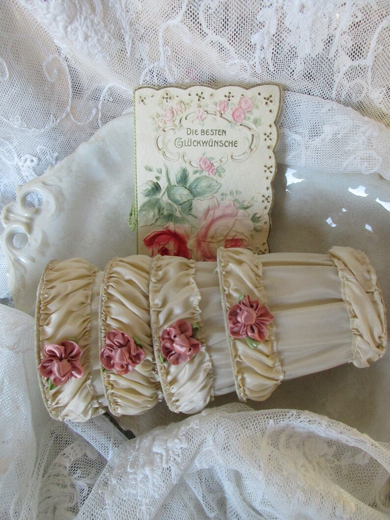May include: Four ivory satin ribbon ruffles with pink satin flowers and green leaves. The ruffles are arranged in a stack and are sitting on a white lace tablecloth. A vintage card with a floral design and the text "Die Besten Gl&uuml;ckw&uuml;nsche" is behind the ruffles.