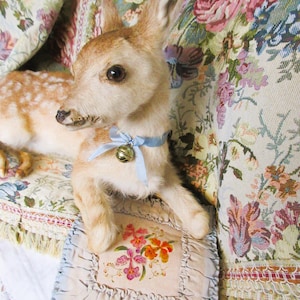 May include: A fawn-colored deer figurine with a light blue ribbon and a small gold bell around its neck. The deer is lying on a floral patterned pillow with a matching floral patterned background.