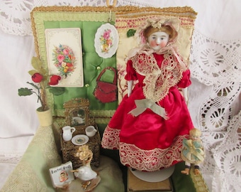 Puede incluir: Una muñeca vintage con un vestido rojo con ribete de encaje y un gorro. La muñeca forma parte de una exhibición en miniatura con un fondo verde, un pequeño espejo y un libro en miniatura. También hay una muñeca pequeña.
