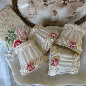 May include: Set of four ivory-colored lampshades with ruffled edges and pink floral accents. The lampshades are arranged on a white surface.
