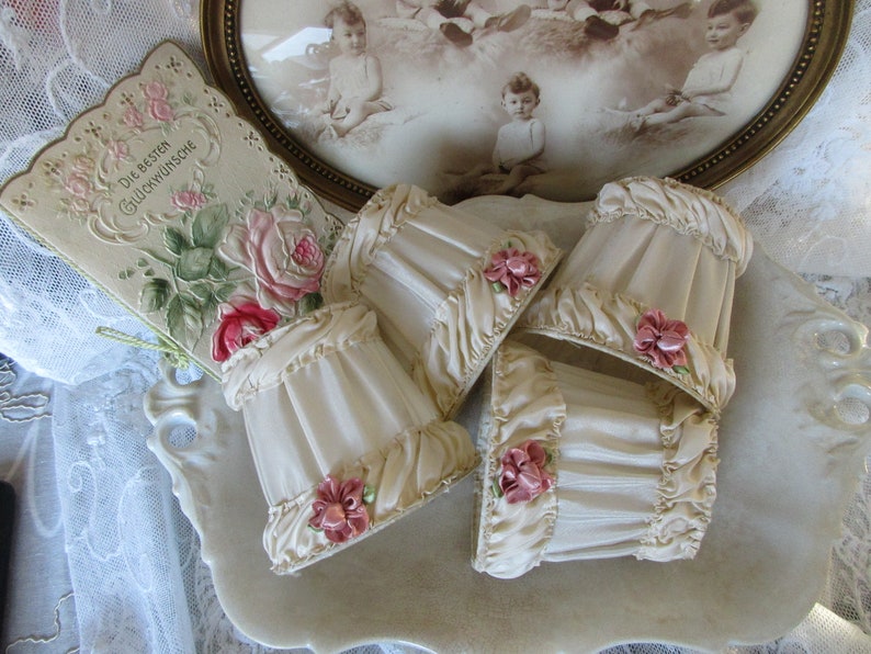May include: Set of four ivory-colored lampshades with ruffled edges and pink floral accents. The lampshades are arranged on a white surface.