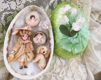 Puede incluir: Un recipiente abierto en forma de huevo contiene una muñeca vintage, un perro pequeño y un botón de querubín. Un segundo huevo verde cubierto de purpurina está decorado con flores blancas y una cinta verde. Los artículos están dispuestos sobre una superficie cubierta de encaje.