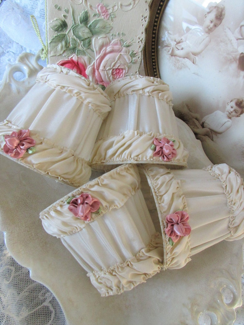 May include: Four ivory-colored lampshades with ruffled edges and pink fabric flowers. The lampshades are arranged on a white surface.
