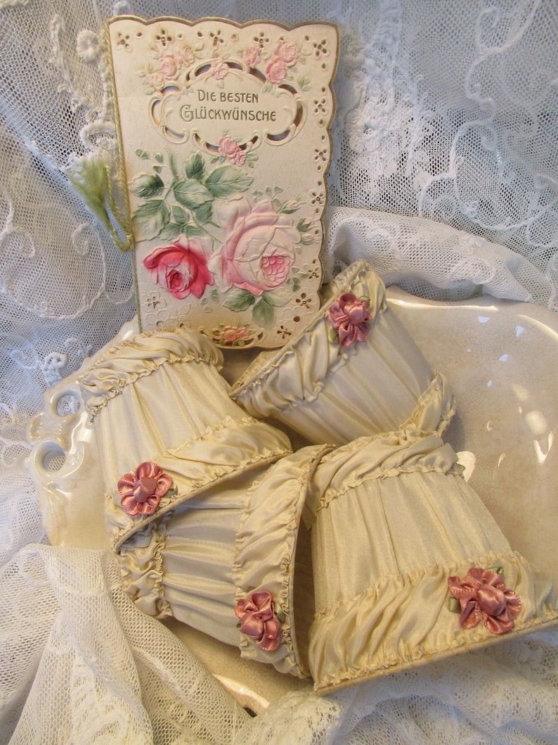 May include: Four cream-colored fabric lampshades with ruffled edges and pink satin bows. The lampshades are arranged on a white porcelain surface with a vintage greeting card featuring a floral design and the text "Die Besten Gl&uuml;ckw&uuml;nsche".