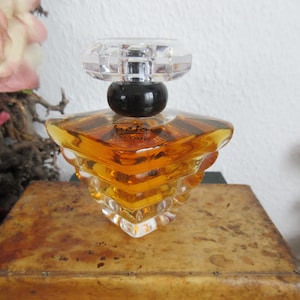 Könnte beinhalten: Ein Parfümflakon aus klarem Glas mit einem facettierten Kristallaufsatz und einem schwarzen kugelförmigen Sockel. Die Flasche hat ein geometrisches Design und enthält eine goldfarbene Flüssigkeit. Das Wort "Tresor" ist auf der Flasche sichtbar.