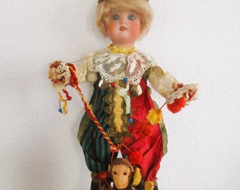 May include: A vintage doll dressed as a jester, wearing a red and gold hat, a green and red outfit, and holding a small monkey on a string. The doll has blonde hair and a painted face. The monkey is brown with red accents.