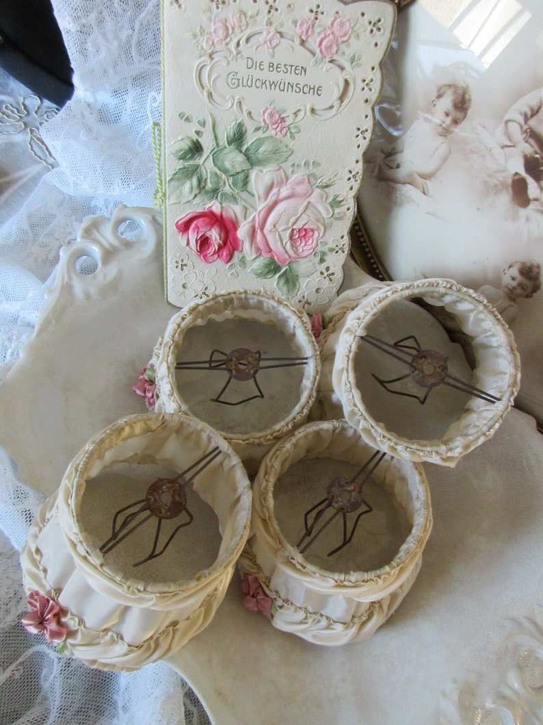 May include: Four vintage lampshades with ruffled edges and delicate pink flowers. The lampshades are made of a cream-colored fabric and have a wire frame.