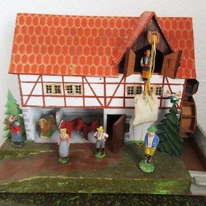 以下が含まれることがあります： 赤い瓦屋根、白と茶色の木骨造りの壁、小さなフィギュアが特徴の、ドイツ風のミニチュア農村風景。 水車、木、人や動物のフィギュアが含まれています。