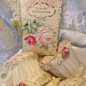 May include: A vintage card with a lace design and floral illustrations. The card reads "Die Besten Gl&uuml;ckw&uuml;nsche" which translates to "Best Wishes". The card is surrounded by four cream-colored fabric lampshades with pink floral embellishments.