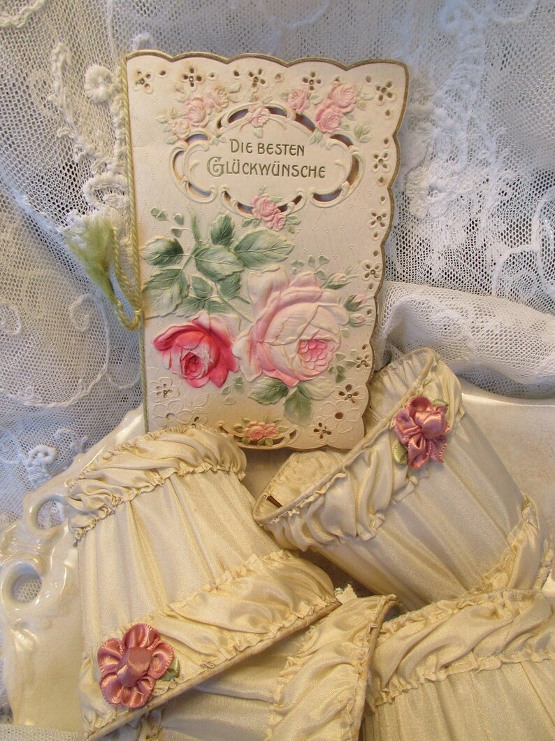 May include: A vintage card with a lace design and floral illustrations. The card reads "Die Besten Gl&uuml;ckw&uuml;nsche" which translates to "Best Wishes". The card is surrounded by four cream-colored fabric lampshades with pink floral embellishments.