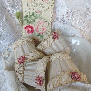 May include: Four cream-colored lampshades with ruffled edges and pink satin bows. The lampshades are sitting on a white lace tablecloth with a vintage card that reads "Die Besten Gl&uuml;ckw&uuml;nsche" in the background.