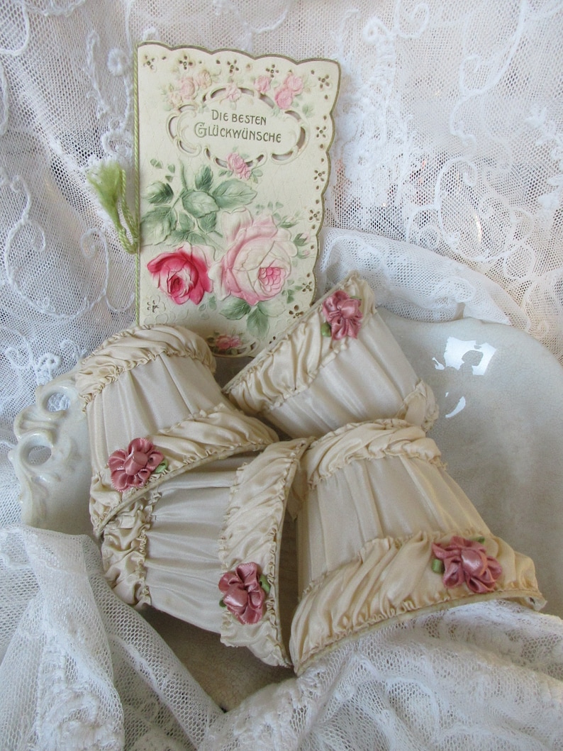 May include: Four cream-colored lampshades with ruffled edges and pink satin bows. The lampshades are sitting on a white lace tablecloth with a vintage card that reads "Die Besten Gl&uuml;ckw&uuml;nsche" in the background.