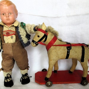 May include: A vintage porcelain doll wearing a brown knitted sweater, braces, and brown trousers, holding a toy rocking horse with red and black details.