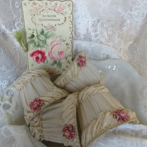 May include: Four cream-colored fabric lampshades with pink satin ribbon rosettes. The lampshades are arranged on a white lace tablecloth with a vintage card featuring a floral design and the text "Die besten Gl&uuml;ckw&uuml;nsche".