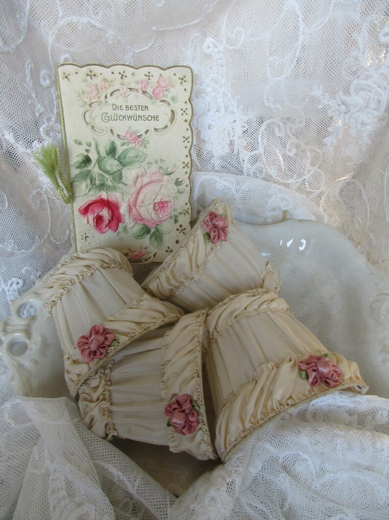 May include: Four cream-colored fabric lampshades with pink satin ribbon rosettes. The lampshades are arranged on a white lace tablecloth with a vintage card featuring a floral design and the text "Die besten Gl&uuml;ckw&uuml;nsche".
