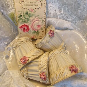 May include: Four cream-colored fabric lampshades with ruffled edges and pink satin bows. The lampshades are arranged in a bowl on a white lace tablecloth. A vintage card with a floral design and the text "Die besten Gl&uuml;ckw&uuml;nsche" is visible in the background.