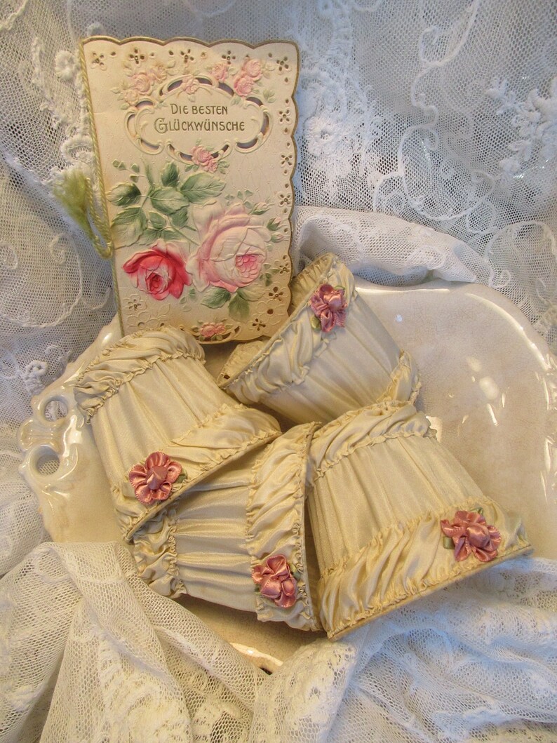 May include: Four cream-colored fabric lampshades with ruffled edges and pink satin bows. The lampshades are arranged in a bowl on a white lace tablecloth. A vintage card with a floral design and the text "Die besten Gl&uuml;ckw&uuml;nsche" is visible in the background.