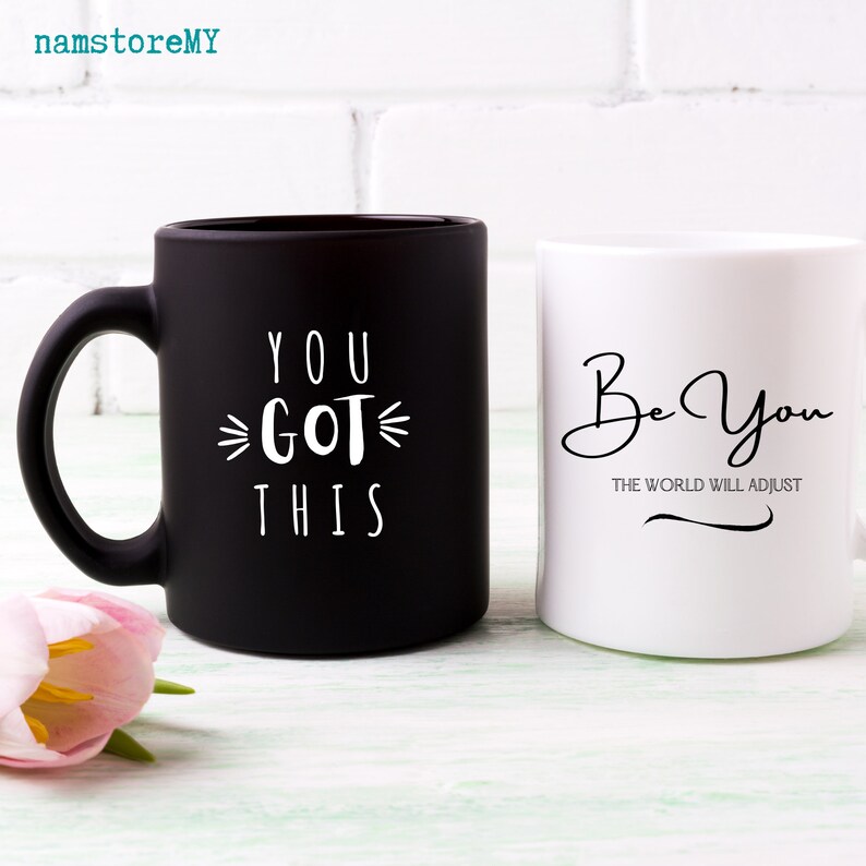 Op de afbeelding: Een matzwarte mok met de witte tekst "YOU GOT THIS" en een glanzend witte mok met de zwarte tekst "Be You THE WORLD WILL ADJUST". Beide mokken hebben een handvat en een cilindrische vorm, gepresenteerd met een roze tulp.