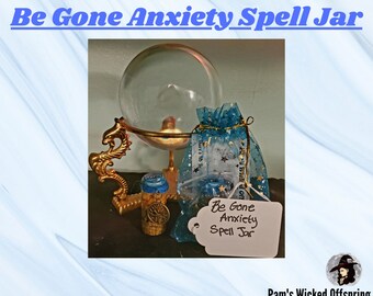 Puede incluir: Un pequeño frasco de vidrio con una tapa azul y plateada y una bolsa de cordón azul y plateada. El frasco está etiquetado como "Be Gone Anxiety Spell Jar". El frasco está sentado en un soporte dorado con una bola de cristal en la parte superior. La imagen tiene un fondo azul y blanco.