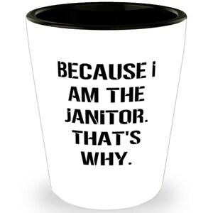 Peut inclure: Verre à shot blanc avec un bord noir, avec le texte "BECAUSE I AM THE JANITOR. THAT'S WHY." en lettres majuscules noires en gras. Le verre a un design conique.