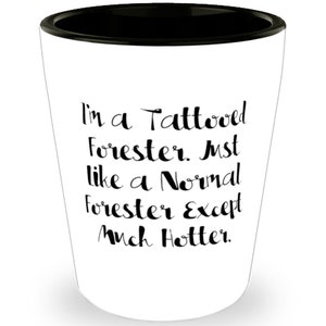 May include: White shot glass with a black rim. The glass has black text that reads "I'm a Tattooed Forester. Just like a Normal Forester Except Much Hotter."