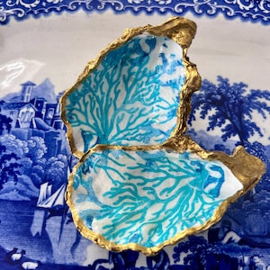 May include: Two decorative oyster shells with gold-painted edges. The shells feature a white interior with a turquoise coral design. The shells are displayed on a blue and white patterned plate.