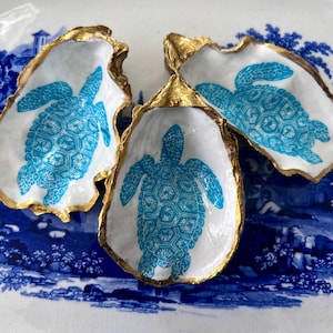 May include: Three oyster shells with gold-painted edges, each featuring a turquoise turtle design on a white background. The shells are arranged on a blue and white decorative plate, creating a coastal theme.