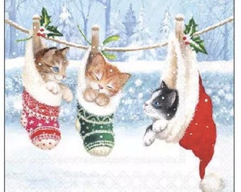 May include: Three kittens in Christmas stockings hanging on a washing line. The stockings are red, green, and red with white trim. The background shows a snowy winter scene with evergreen trees.