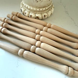 May include: A collection of unfinished wooden spindles, likely for furniture or decorative projects. The spindles are light-colored, with a turned design featuring rounded sections and a smooth finish. A decorative white base is in the background.