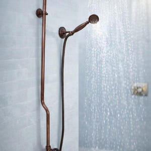 May include: A wall-mounted shower system in an oil-rubbed bronze finish featuring a large circular rainfall showerhead, a handheld shower wand on a flexible hose, and a dual-handle faucet control. Water flows from the rainfall showerhead.
